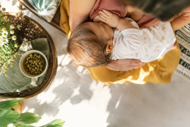 Une mère allaite son bébé dans un environnement chaleureux avec une tisane aux fleurs posée sur une table à proximité.