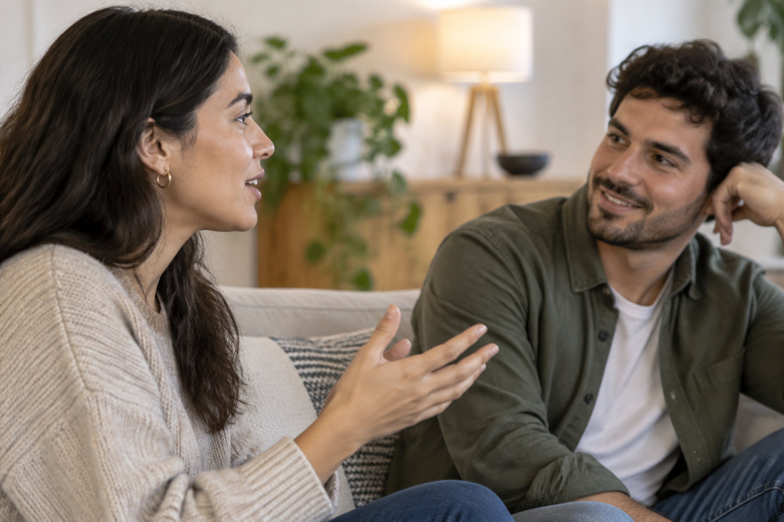 Une femme gesticule en parlant à un homme qui l’écoute avec un sourire, assis sur un canapé dans un salon.